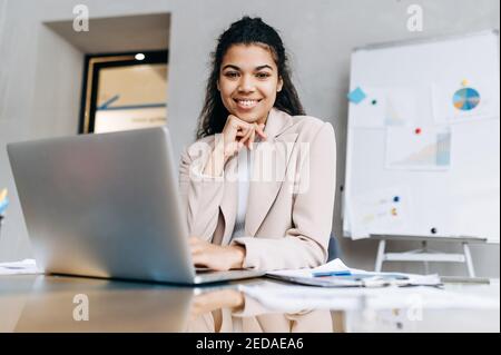 Une femme d'affaires élégante s'assoit au bureau, souriant et regarde la caméra. Une femme afro-américaine utilisant un ordinateur portable développe un nouveau projet. Belle jeune femme adulte étudiant ou travaillant à distance Banque D'Images