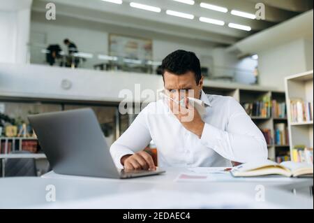 Homme d'affaires hispanique éternuant, utilisant un papier de soie, se sentant mal. Gestionnaire malsain qui lutte avec le nez courant, attraper une grippe, travailler à distance ou étudier, pendant la maladie Banque D'Images