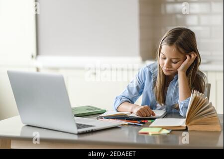 La jeune fille est fatiguée de la leçon en ligne. Apprentissage à distance pendant la quarantaine. Focalisé caucasien jolie écolière faisant ses devoirs à la maison à l'aide d'un ordinateur portable, assis au bureau Banque D'Images