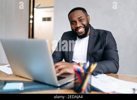Un bon Manager ou patron est assis au bureau. Homme d'affaires afro-américain utilisant un ordinateur portable, développer un nouveau projet ou démarrer. Un employé plein de confiance regarde la caméra, souriant Banque D'Images