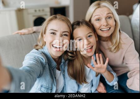 Trois générations de femmes. Gaie grand-mère caucasienne, fille et petite-fille sont assis à la maison sur le canapé, embrassant, prend selfie et souriant. Relations familiales et valeurs Banque D'Images