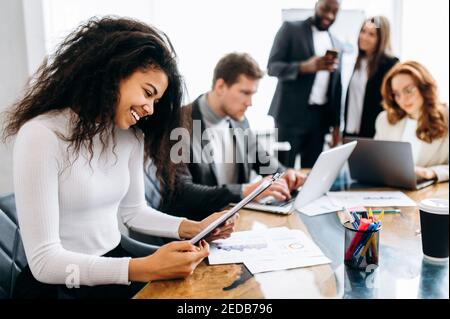 Femme afro-américaine réussie à la réunion d'affaires. Belle femme employeur, l'apprentissage de graphiques financiers, faire de la paperasserie, sourire. Jeune adulte gérant assis au bureau dans un bureau moderne Banque D'Images