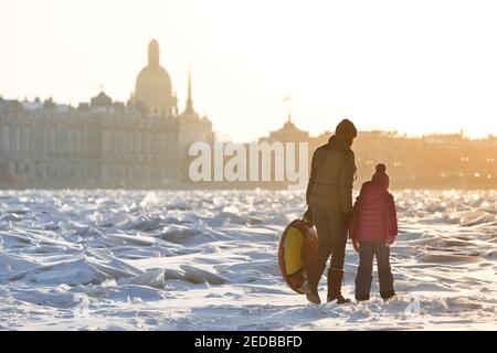 Maman et enfant marchant sur la glace de la rivière gelée en hiver ensoleillé, paysage urbain en arrière-plan Banque D'Images