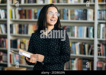 Portrait d'une jeune femme d'affaires freelance ou étudiante en chemise formelle et lunettes debout dans un bureau moderne, tenir dans les bras des documents et des livres, regarde loin et sourire Banque D'Images