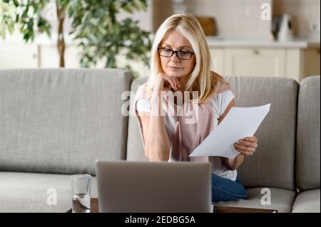 Un blond senior ciblé travaille à distance à la maison, assis sur le canapé dans le salon, analyse les rapports mensuels, utilise un ordinateur portable. Travailler à domicile Banque D'Images