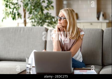 Blond senior frustré fatigué des travaux en ligne, assis sur le canapé dans le salon, utilise un ordinateur portable, regarder loin, besoin d'un repos, rêve de vacances ou de week-end Banque D'Images