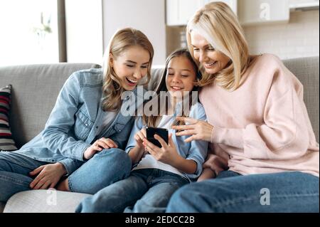 Gaie grand-mère caucasienne, fille et petite-fille passant du temps ensemble, assis à la maison sur le canapé à l'aide d'un smartphone, de naviguer sur le réseau, de faire des achats en ligne, prend selfie, souriant Banque D'Images