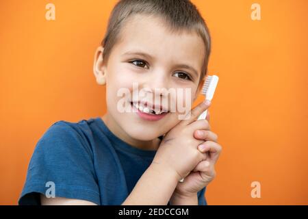 Un garçon sans la dent supérieure de lait dans un t-shirt bleu tient la brosse à dents à la main sur fond orange. Banque D'Images