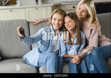 Trois générations. Une grand-mère, une fille et une petite-fille du caucase souriant et heureux, assis à la maison sur le canapé, embrassant, prend le selfie et souriant. Relations familiales et valeurs Banque D'Images