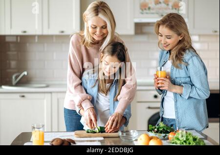 Trois générations de femmes passent du temps ensemble dans la cuisine, la grand-mère enseigne à la petite-fille de couper la salade et les montres de maman souriant et de boire du jus à côté d'eux Banque D'Images