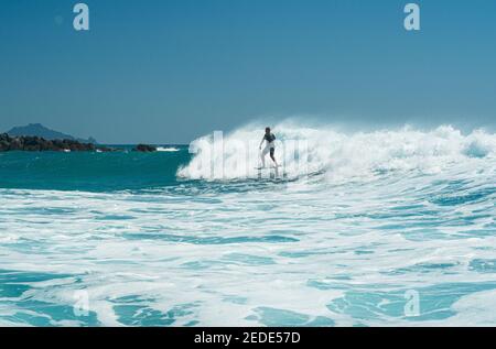 Un homme surfe sur une planche de surf hydrofoil sur des vagues turquoise à Mangawhai Heads, en Nouvelle-Zélande. Ciel bleu clair. Banque D'Images