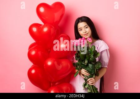 Concept de la Saint-Valentin. Jeune fille tendre et douce tenant des fleurs romantiques, debout près des ballons rouges de coeur de l'amant et regardant avec l'amour Banque D'Images