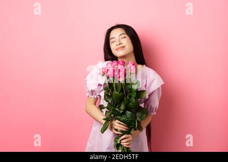 Jeune fille asiatique rêveuse se sentant romantique, tenant des fleurs et rêvant, l'imagerie Saint Valentin date, portant une jolie robe, debout sur fond rose Banque D'Images