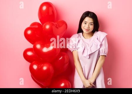 Jolie fille asiatique en robe regardant timide et souriant, debout modeste près des ballons de jour de Saint Valentin, rougir sur la date romantique, regardant l'appareil photo, rose Banque D'Images