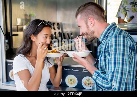 Doux couple interracial regardant dans les yeux de l'autre tout en mangeant des pâtes dans un camion alimentaire Banque D'Images