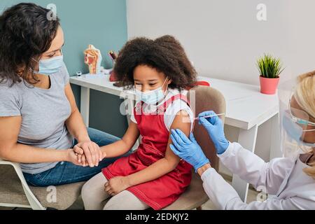 Vaccinations de routine pour un enfant. Une fille afro-américaine et sa mère portent un masque médical lorsqu'elles sont vaccinées par un médecin généraliste Banque D'Images