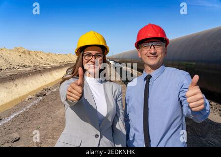 Homme et femme souriants dans des casques de sécurité avec des Thumbs vers le haut au site de construction de gazoduc. Des gens d'affaires prospères dans Hardhats debout à côté de Pipeline. Banque D'Images