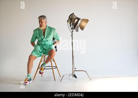 Photo studio d'un homme d'âge moyen à cheveux gris tendance portant une tenue élégante avec des baskets colorées à côté, assis à côté du spot du studio, posé sur fond blanc. Mode, concept de style Banque D'Images