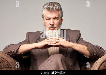 Portrait d'un homme d'affaires brutal et mature avec une barbe vêtue d'un élégant costume regardant l'appareil photo, assis avec les mains croisées dans un fauteuil tout en posant isolé sur un arrière-plan clair. Vue avant Banque D'Images