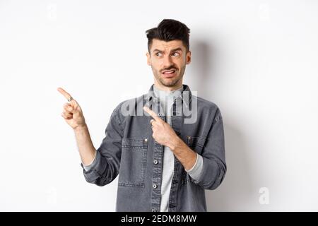 Jeune homme sceptique et douteux pointant les doigts à gauche sur le logo, regarde avec déplaît et confusion, debout sur fond blanc Banque D'Images