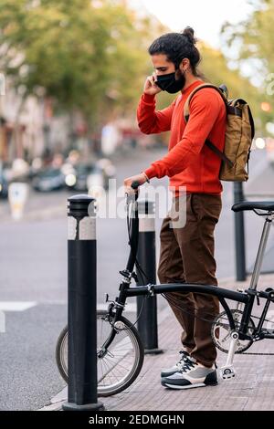 Photo de stock de l'homme portant un masque facial dû à covid19 utilisant son téléphone dans la rue et tenant un vélo. Banque D'Images