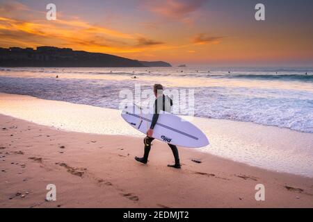 Coucher de soleil sur un surfeur transportant sa planche de surf et marche le long du rivage sur la plage de Fistral à Newquay, en Cornouailles. Banque D'Images