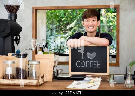 Jeune homme asiatique joyeux entrepreneur au comptoir dans son propre café avec le signe du café J'aime Banque D'Images