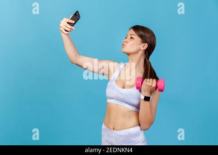 Profil portrait femme mince blogueur en blanc sportswear posant sur l'appareil photo de son smartphone, enregistrement de l'entraînement avec dumbbell tutorial. Salle de studio intérieure Banque D'Images