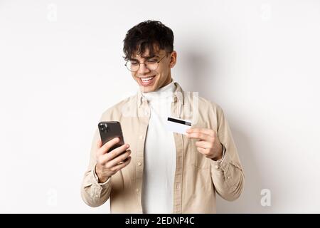 Achats en ligne. Jeune homme heureux utilisant un smartphone et une carte de crédit en plastique, payant sur Internet, debout sur fond blanc Banque D'Images