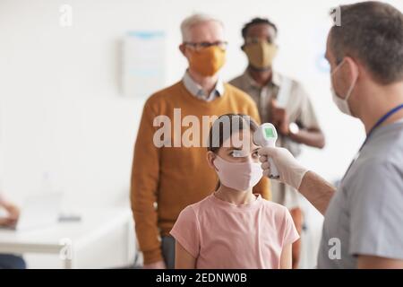 Prise de vue d'un médecin méconnaissable vérifiant la température d'une petite fille portant un masque et attendant en file d'attente à la clinique ou à l'hôpital, espace de copie Banque D'Images