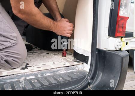 Homme barbu perçant un trou avec un outil de batterie dans le sol d'un véhicule. Auto-construction d'une camionnette de camping dans la rue. Banque D'Images