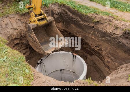 Pelle hydraulique avec un godet, s'abaissant dans la fosse sur des câbles en acier anneau d'égout en béton. Construction ou réparation d'un égout. Banque D'Images