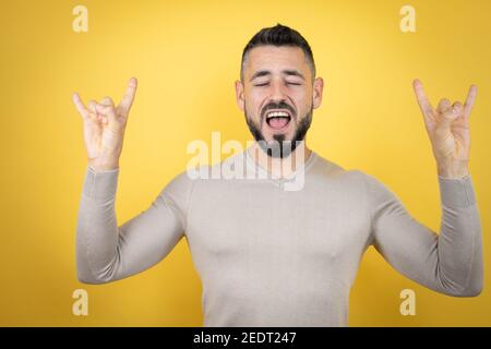 Beau homme avec barbe portant un pull sur fond jaune criant avec une expression folle faisant le symbole rock avec les mains vers le haut Banque D'Images
