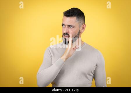 Beau homme avec la barbe portant un pull sur fond jaune main sur la bouche en disant secret rumeur, chuchotant la conversation malveillante Banque D'Images