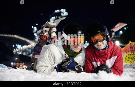 Couple de skieurs allongé sur une pente enneigée sous un télésiège éclairé la nuit, se reposant après avoir passé du temps actif sur les pistes de ski. Portrait en gros plan de deux heureux joyeux sur la neige Banque D'Images