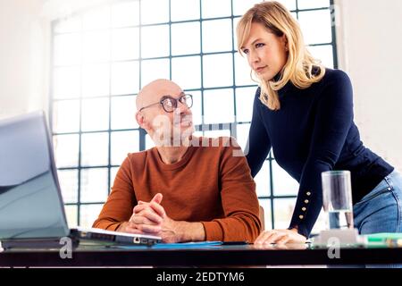 Groupe de gens d'affaires occasionnels travaillant ensemble au bureau. Homme d'affaires assis à un bureau derrière son ordinateur portable tandis qu'une femme d'affaires se tient à côté de lui Banque D'Images