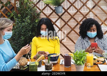 Amis multiraciaux utilisant un smartphone mobile tout en ayant un déjeuner sain à l'intérieur café brunch bar pendant l'épidémie de virus corona - nourriture et concept de technologie Banque D'Images