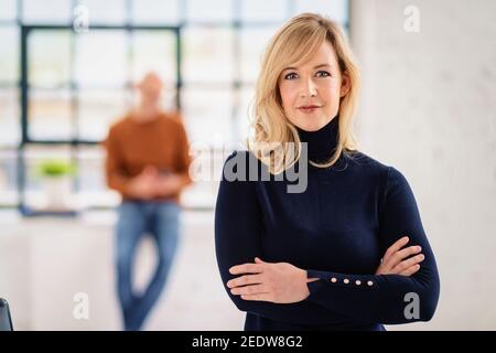Portrait tiré belle femme blonde aux cheveux debout avec les bras croisés dans le salon tandis que l'homme méconnaissable debout dans l'arrière-plan. Banque D'Images