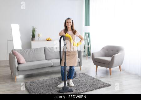 Service de ménage. Bonne jeune femme de ménage avec aspirateur au milieu d'un salon bien rangé, portrait en longueur Banque D'Images