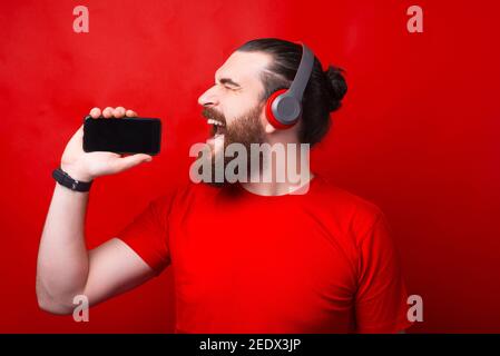 Jeune barbu tenant un smartphone et signant et écoutant de la musique sur un casque sur fond rouge. Banque D'Images