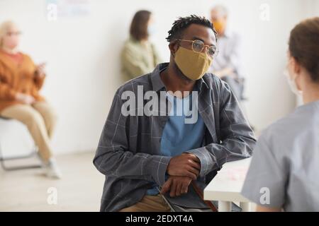 Portrait du jeune Africain américain homme portant le masque tout en parlant au médecin ou à l'infirmière dans la clinique médicale, l'espace de copie Banque D'Images