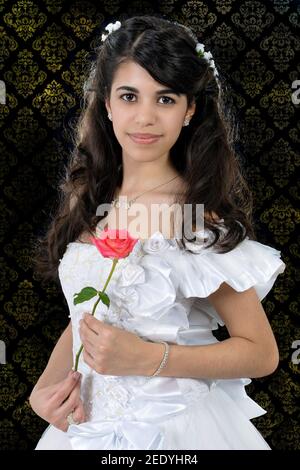 Portrait de seize ou de quinces doux: Belle fille latine portant une robe blanche avec de longs cheveux noirs et tenant une rose rose Banque D'Images