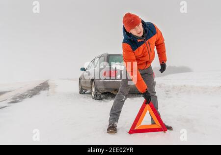 Homme dans une veste orange place un triangle d'avertissement sur une route enneigée contre le fond d'une voiture. Concept de panne de voiture, accident Banque D'Images