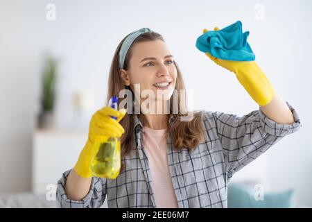 Belle femme millénaire dans des gants en caoutchouc essuyant la fenêtre avec le spray détergent à la maison Banque D'Images