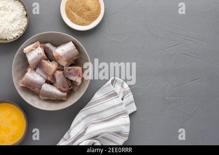 Dans un bol, des morceaux de merlu et des produits pour la cuisine frite, des œufs, de la farine et de la chapelure ont été présentés avec une serviette de cuisine sur fond gris Banque D'Images