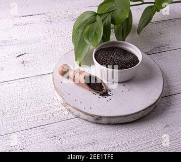 Graines de quinoa brutes noires dans un bol blanc et une cuillère en bois avec des graines sur une table en bois blanc. Peu de graines sont dispersées autour. Feuilles vertes à l'arrière-plan. Il Banque D'Images