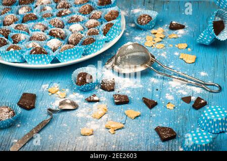 Boule de chocolat dans un panier de papier à pois bleu-blanc sur une assiette blanche sur un plan de cuisine bleu avec sucre renversé, biscuit et morceau de chocolat, passoire et cuillère. Banque D'Images