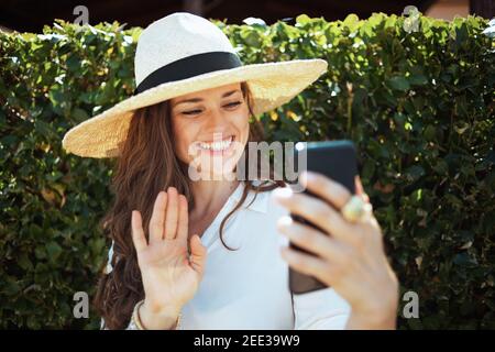 jolie femme élégante en chemise blanche avec chapeau ayant une réunion vidéo sur un smartphone à l'extérieur près du mur vert. Banque D'Images