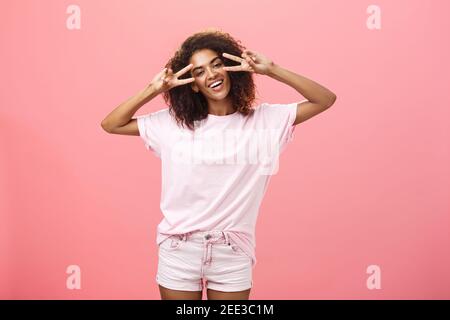 Photo en intérieur d'une femme à la peau sombre et charismatique avec une coiffure afro tête inclinable montrant des signes de paix ou de victoire sur les yeux et sourire Banque D'Images