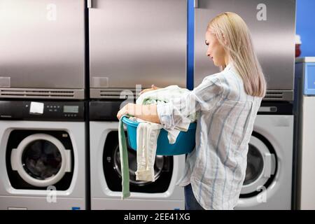 vue arrière sur la femme est venu de mettre des vêtements à laver dans la maison de lavage, attendant seul, tenant lavabo avec énorme pile de choses Banque D'Images